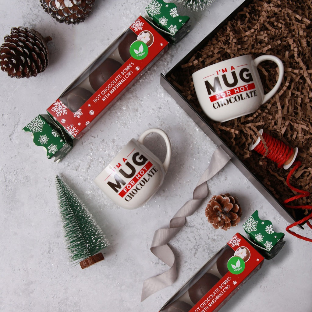 Two Hot Chocolate mugs with Christmas Crackers containing hot chocolate bombes, on a decorative snowy background