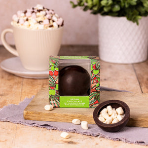 Vegan dark hot chocolate bombe with vegan marshmallows, showing in front of a mug of delicious hot chocolate with vegan cream and vegan marshmallows