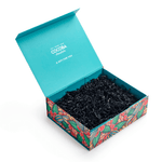 Open teal box with colorful floral design containing black chocolate pieces on a white background